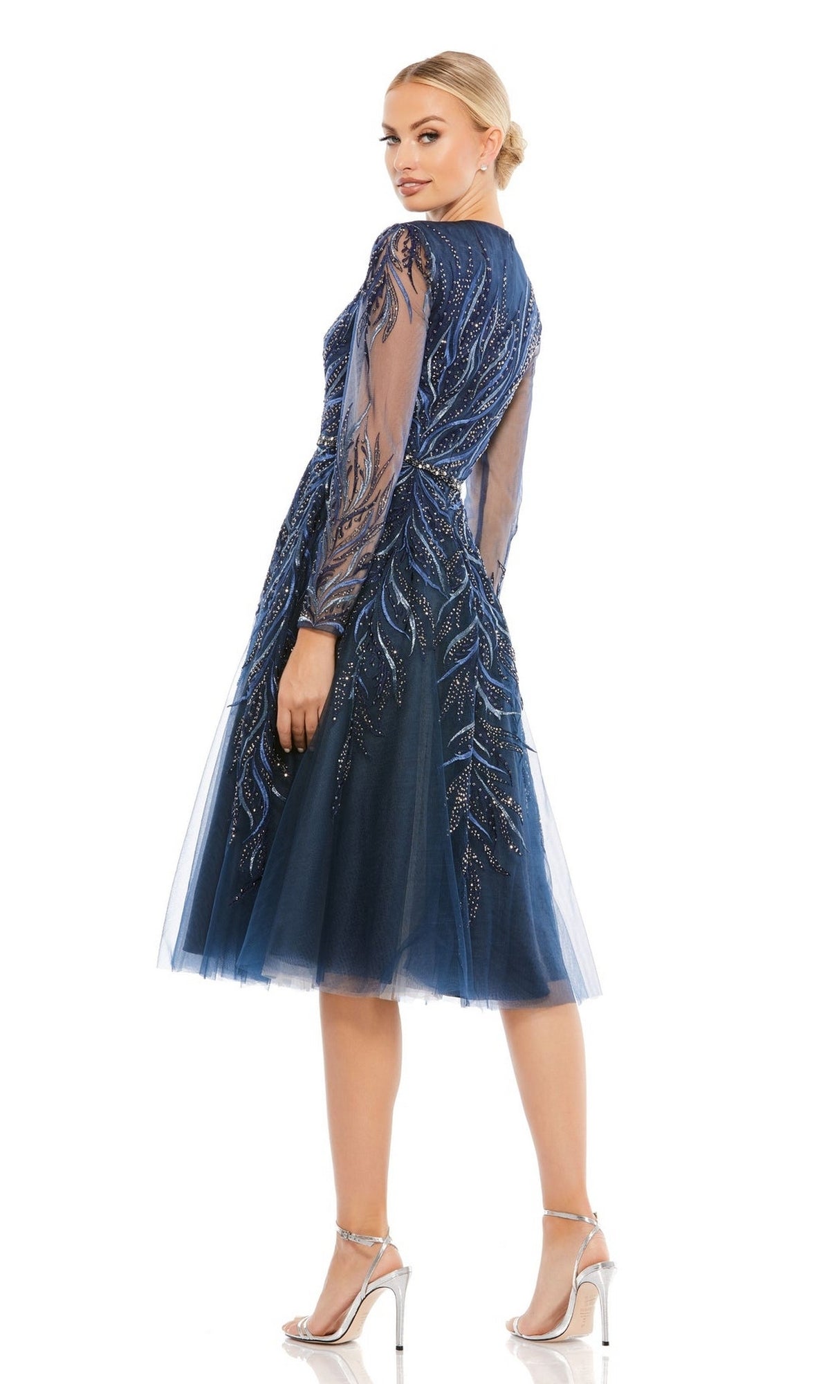 A woman in the Mac Duggal 11136 blue knee-length cocktail dress with sheer long sleeves, sequin detailing, and silver high heels stands facing slightly away.